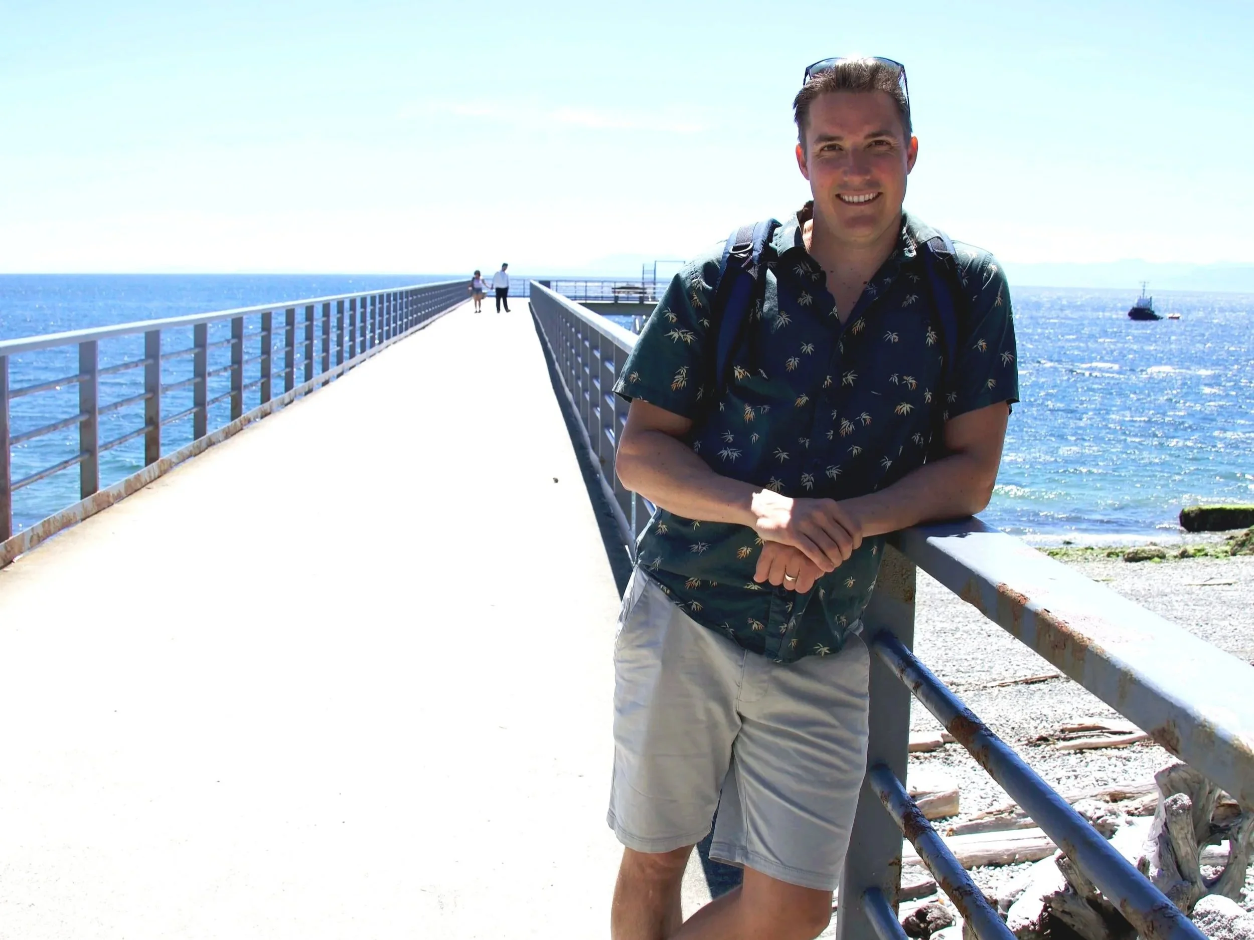 A man standing on a pier by the ocean, smiling at the camera, wearing a patterned short-sleeve shirt and shorts, with glasses on his head, with two people walking in the background and a boat on the water.