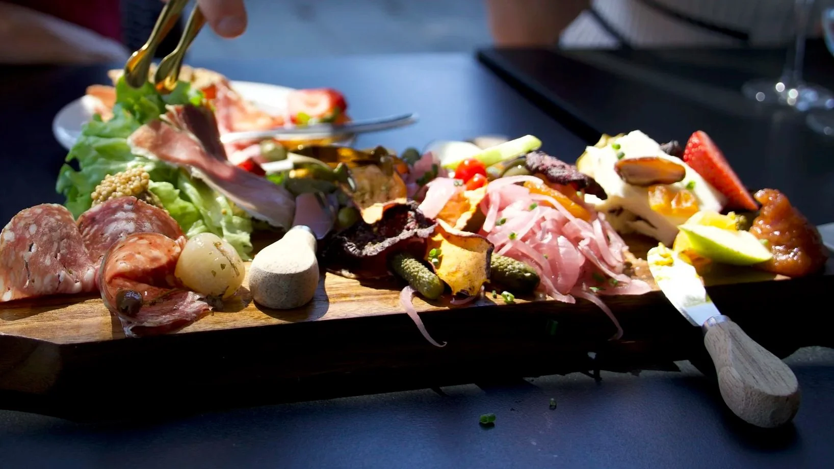 A wooden charcuterie board with various meats, cheeses, pickles, and garnishes, with someone using tongs to serve.