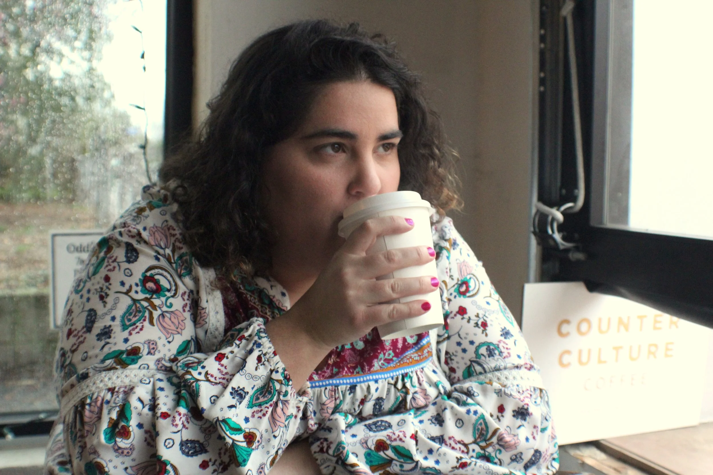Woman with dark, curly hair drinking from a white disposable cup inside a building, wearing a colorful, patterned blouse.