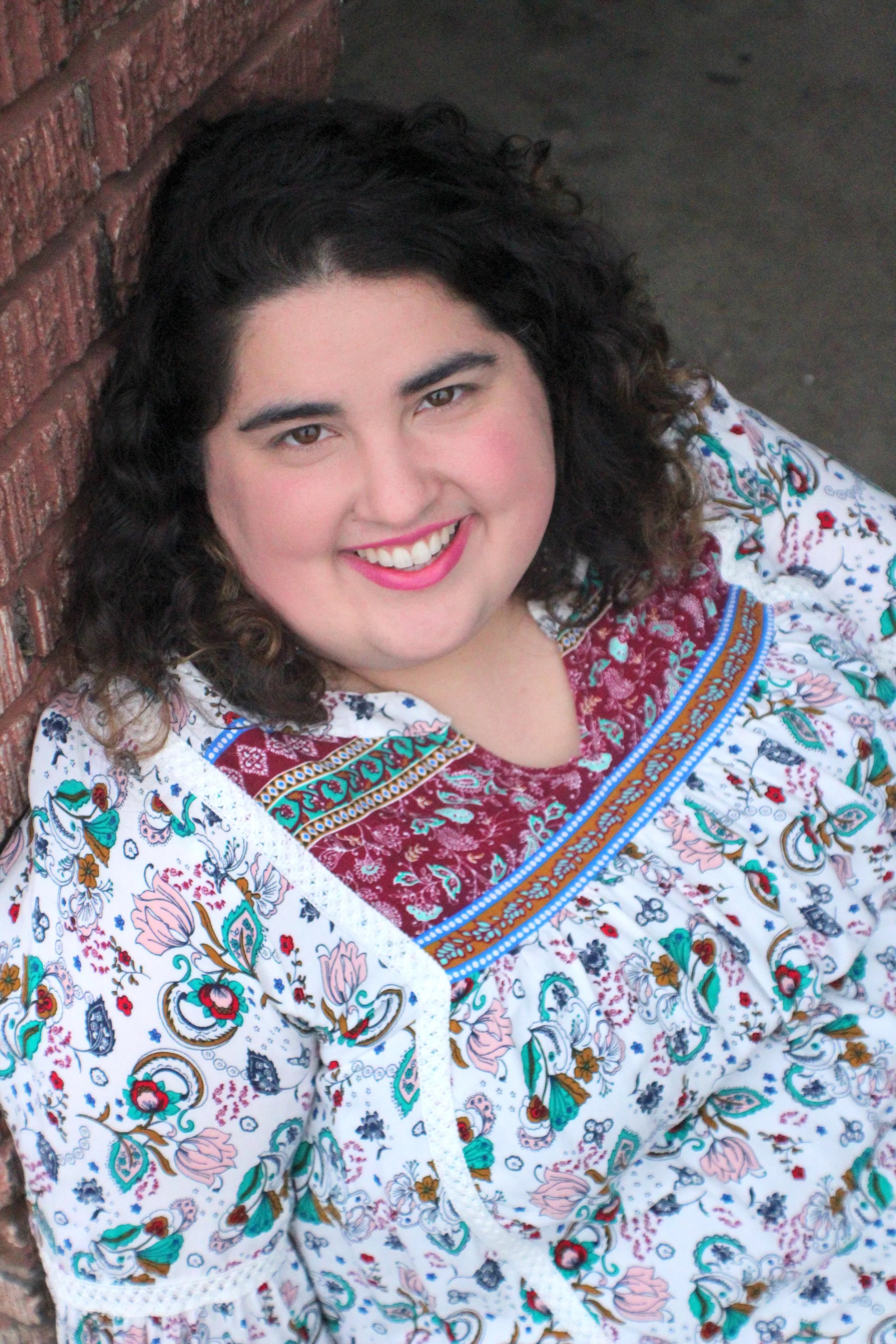 Close-up of a woman with dark curly hair, wearing makeup with rosy cheeks and pink lipstick, smiling, and dressed in a colorful, patterned blouse with floral and paisley designs, leaning against a brick wall.