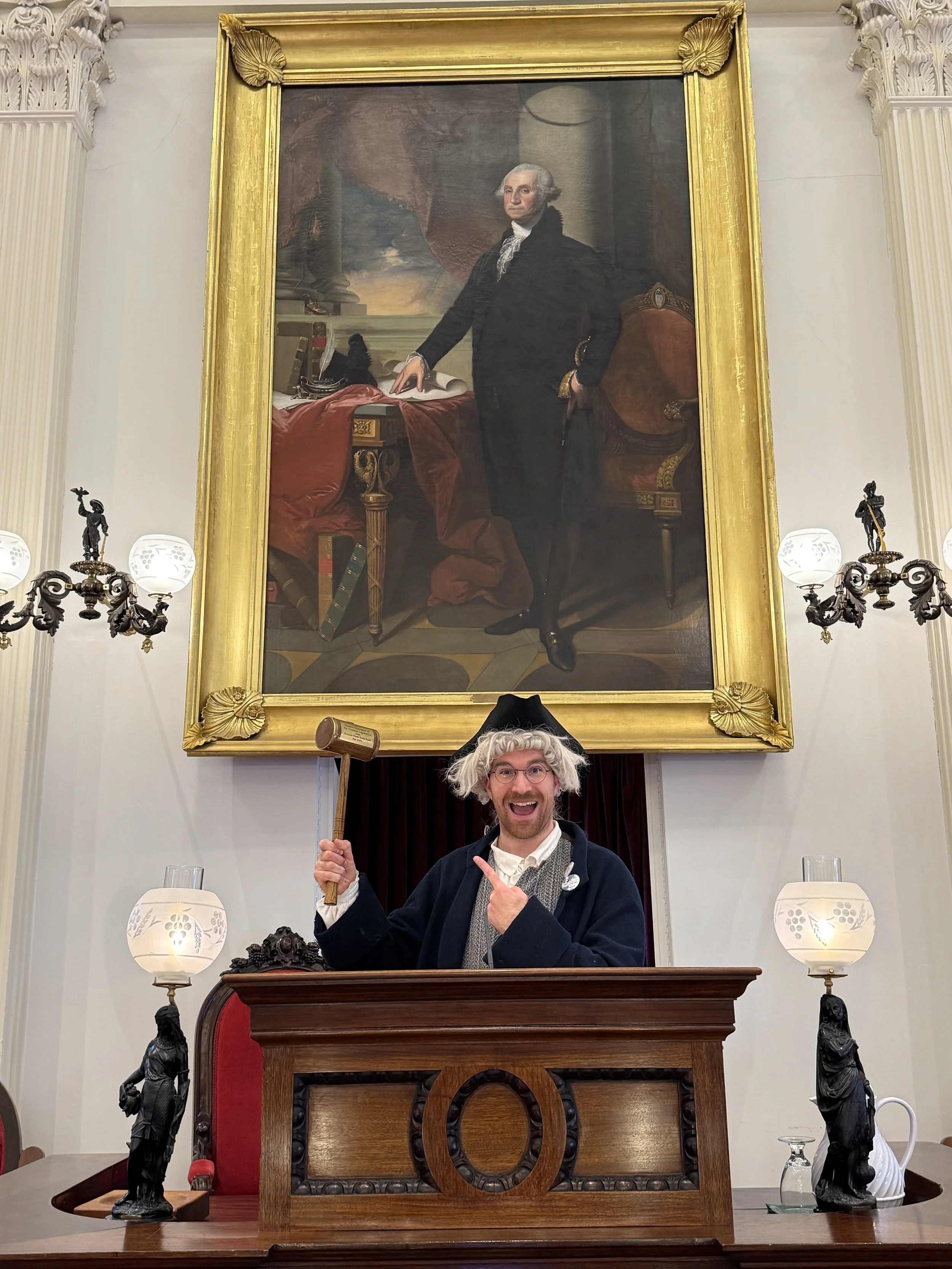 A person dressed as George Washington, wearing a wig, standing behind a wooden desk in a room with large portrait painting of George Washington hanging above. The person is smiling and holding a gavel, surrounded by ornate lamps and statues of colonial figures.
