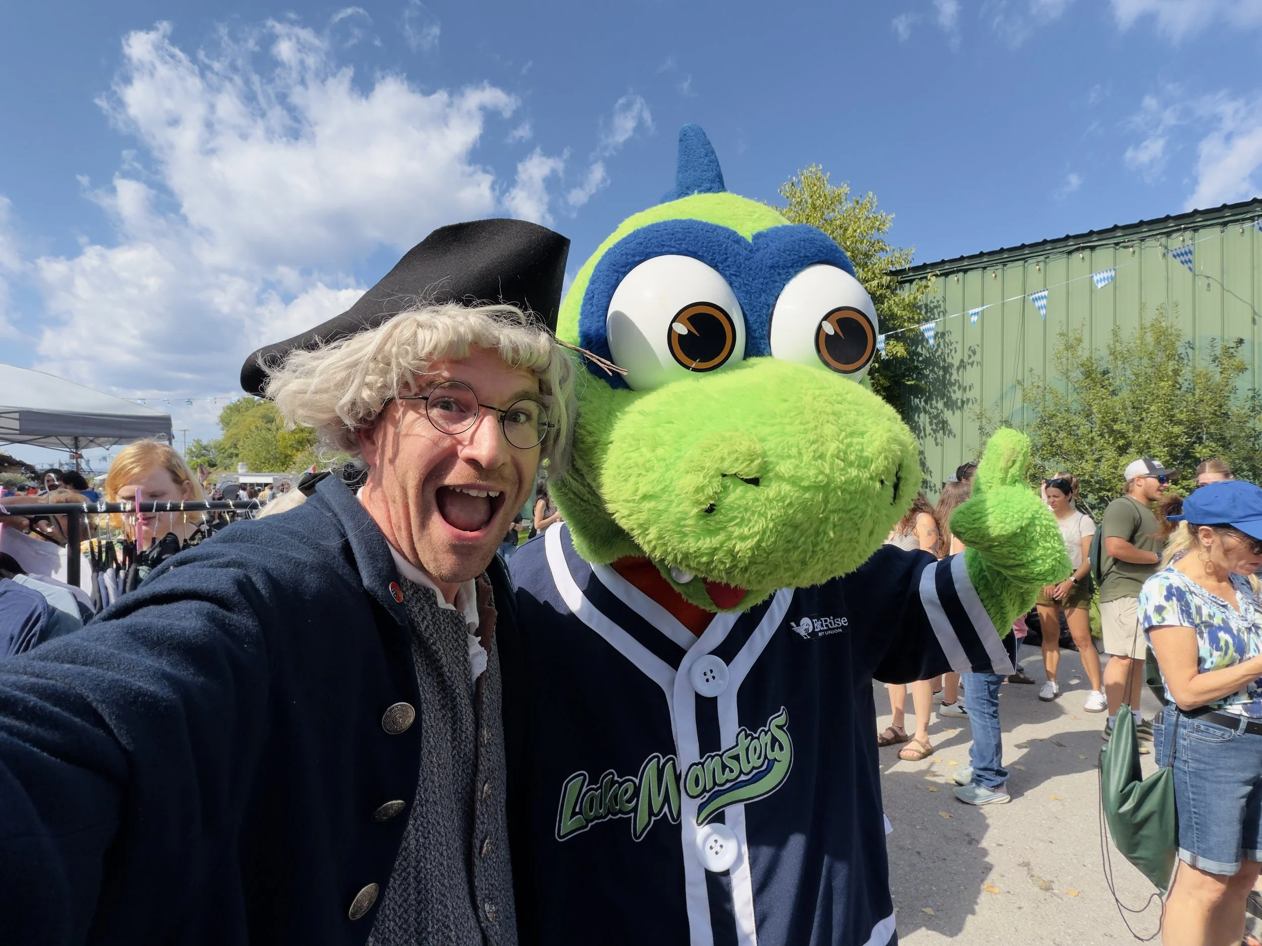 A person with curly blonde hair, glasses, and a black jacket taking a selfie next to a person in a large, green and blue dinosaur costume at an outdoor event with a crowd and blue sky in the background.