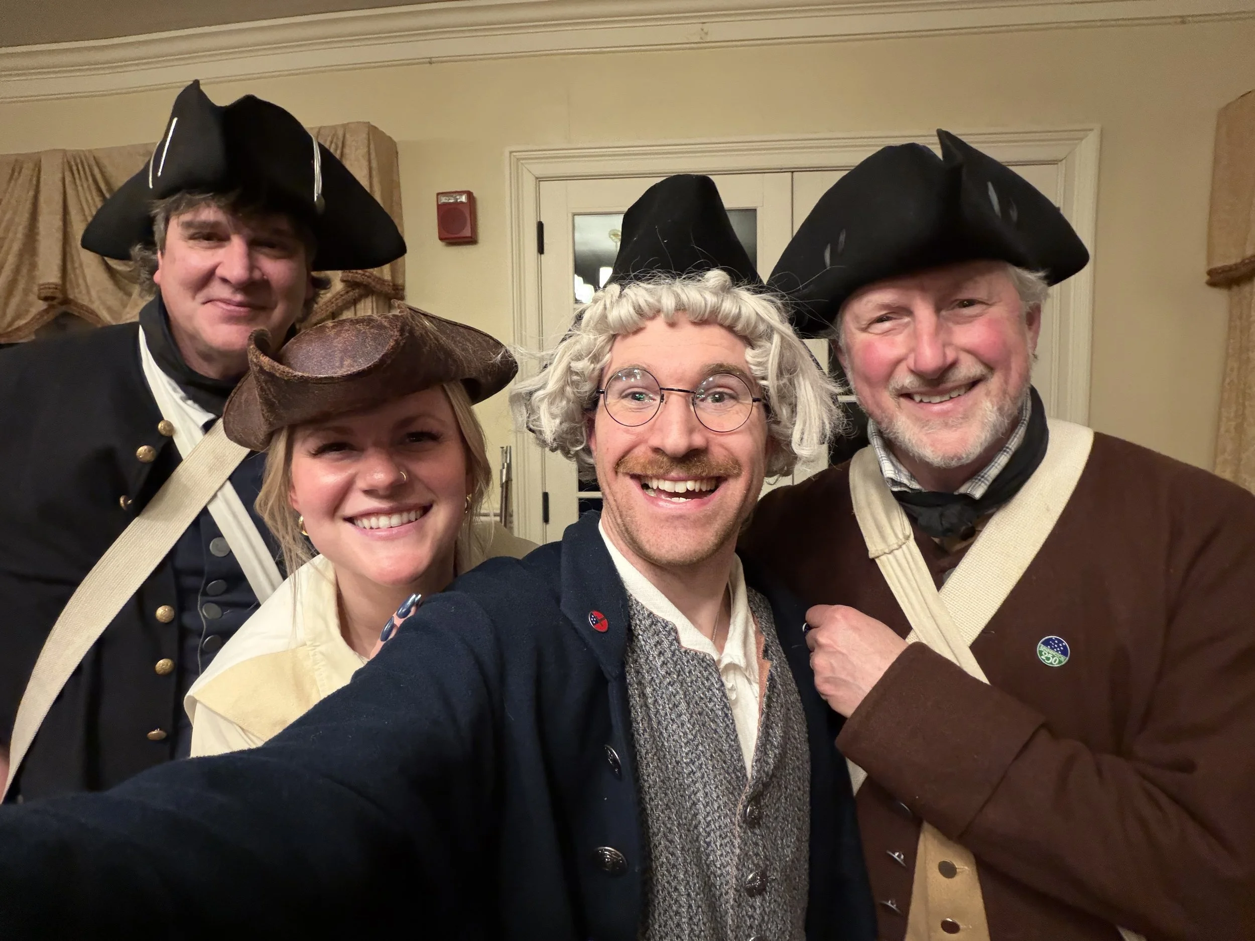 Group of four adults dressed in historical costumes, wearing tricorn hats, and smiling for the camera inside a room with beige walls and curtains.