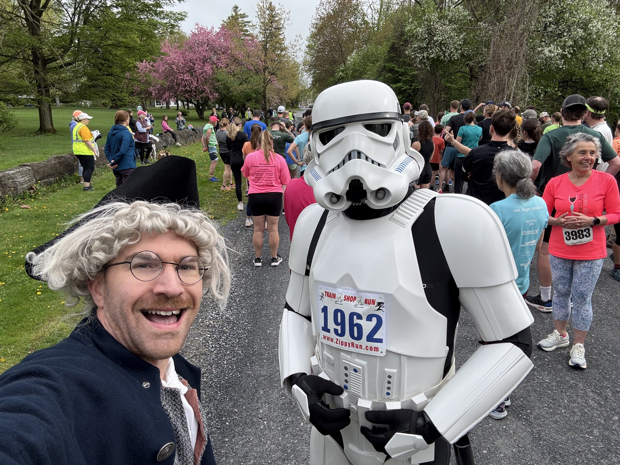 A man smiling and taking a selfie with a crowd of people gathered for a race in the background. The crowd includes a person in a Stormtrooper costume. Trees and pink blooming flowers are visible in the park area.