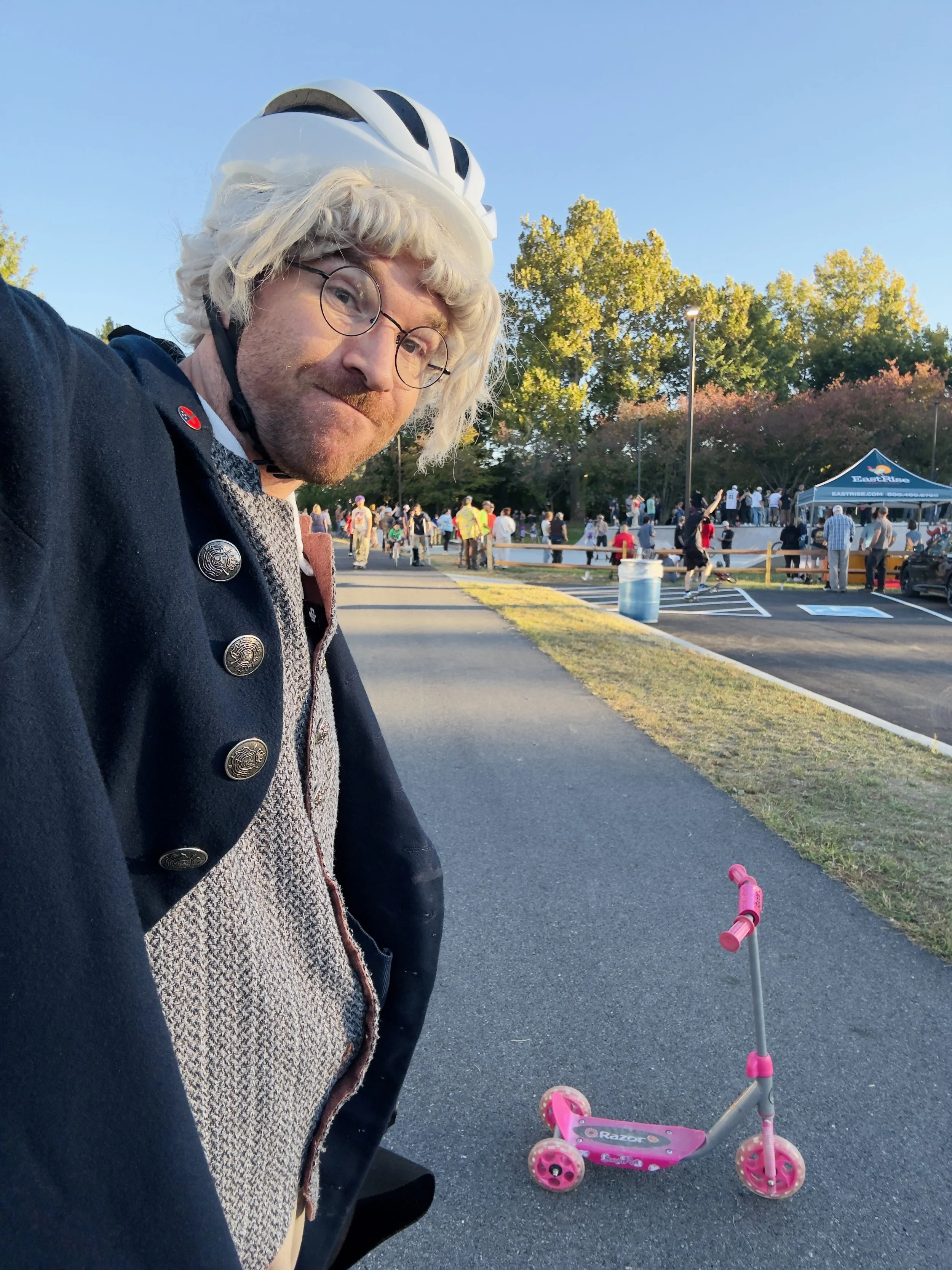 A man with blonde curly hair, glasses, and a beard taking a selfie at a park during daytime. He is wearing a helmet and a navy jacket, and there is a pink children's scooter on the ground nearby. Behind him, people are gathered around a skate park.
