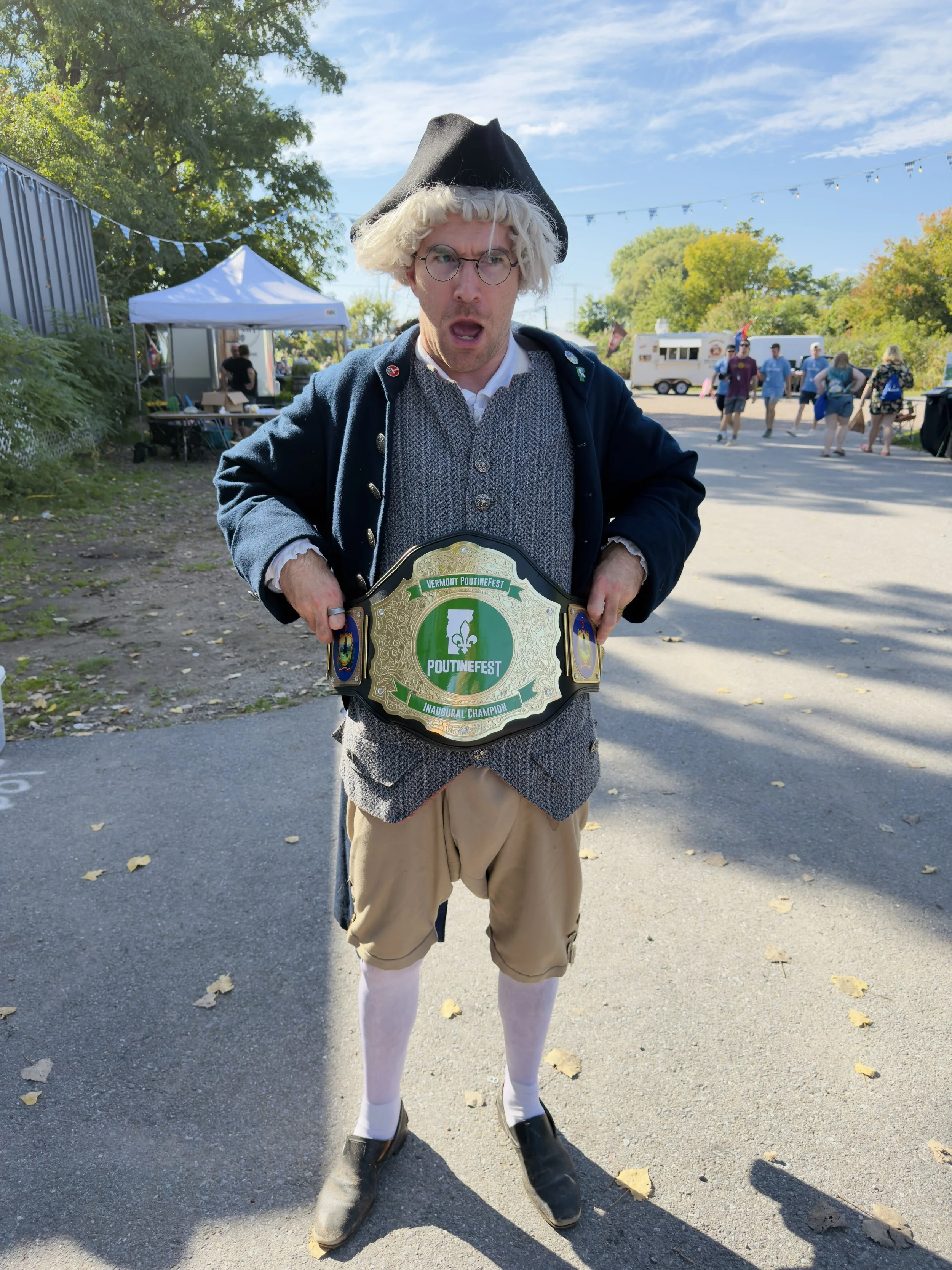 Man dressed as a humorous version of a historical character, wearing a black tricorn hat, round glasses, a buttoned coat, a vest, and beige shorts with white tights, standing outdoors at an event with a championship belt reading 'Vermont PoutineFest Inaugural Champion.'
