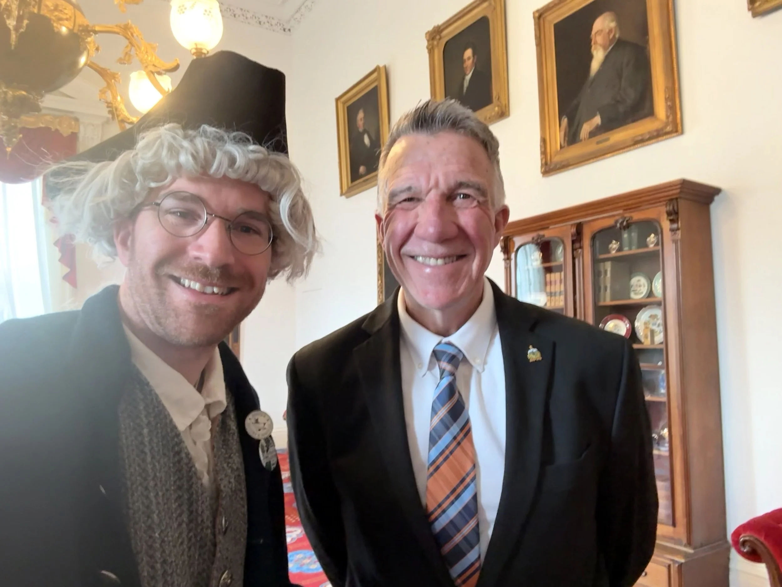 Two men smiling and taking a selfie in a room with framed portraits on the wall, a wooden display cabinet, and ornate gold and white decor.