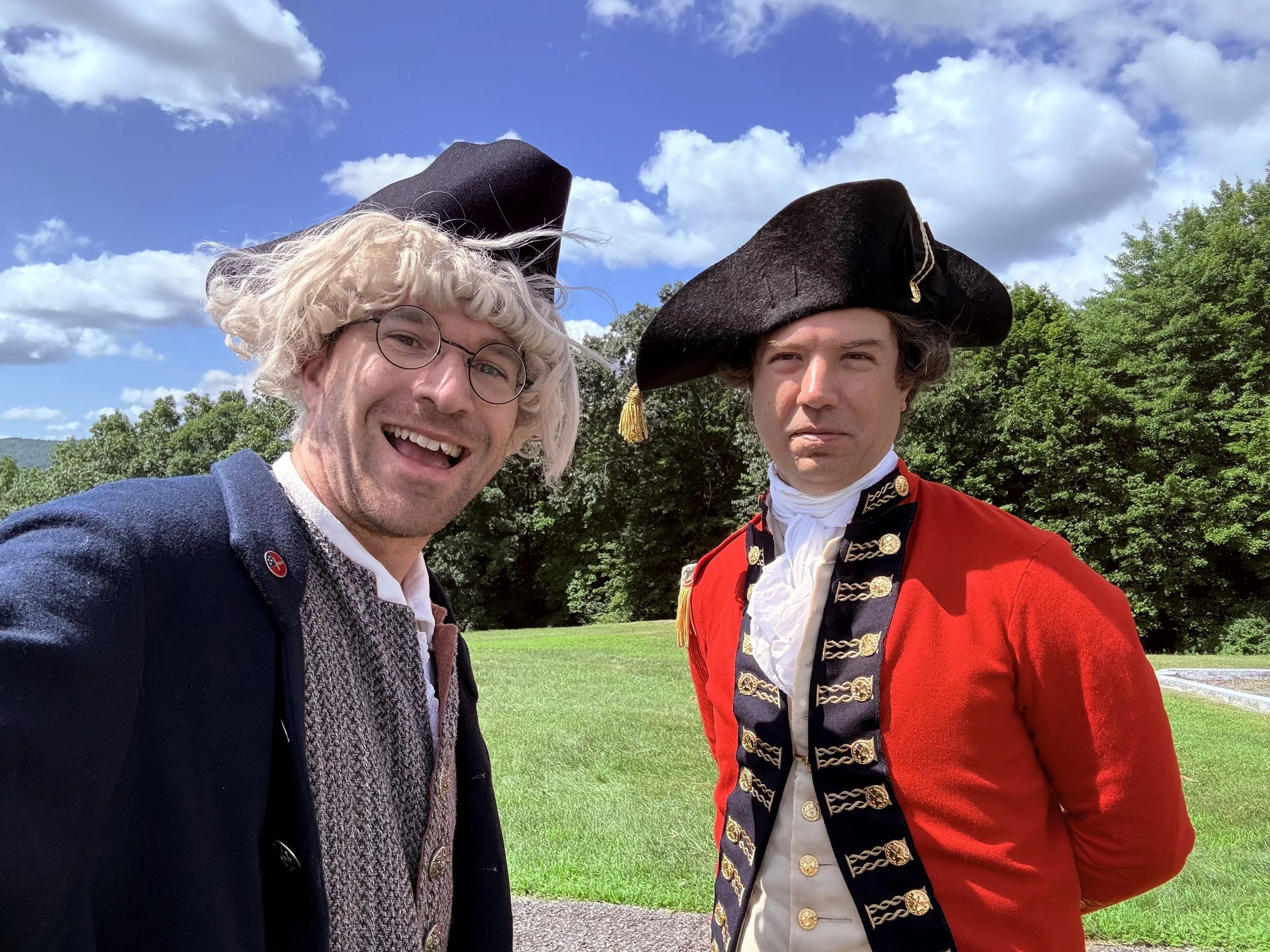 Two men taking a selfie outdoors, one dressed as a historical figure in a red coat with gold accents and a tricorn hat, the other in modern attire with curly blond hair and round glasses, against a background of green trees and blue sky with white clouds.