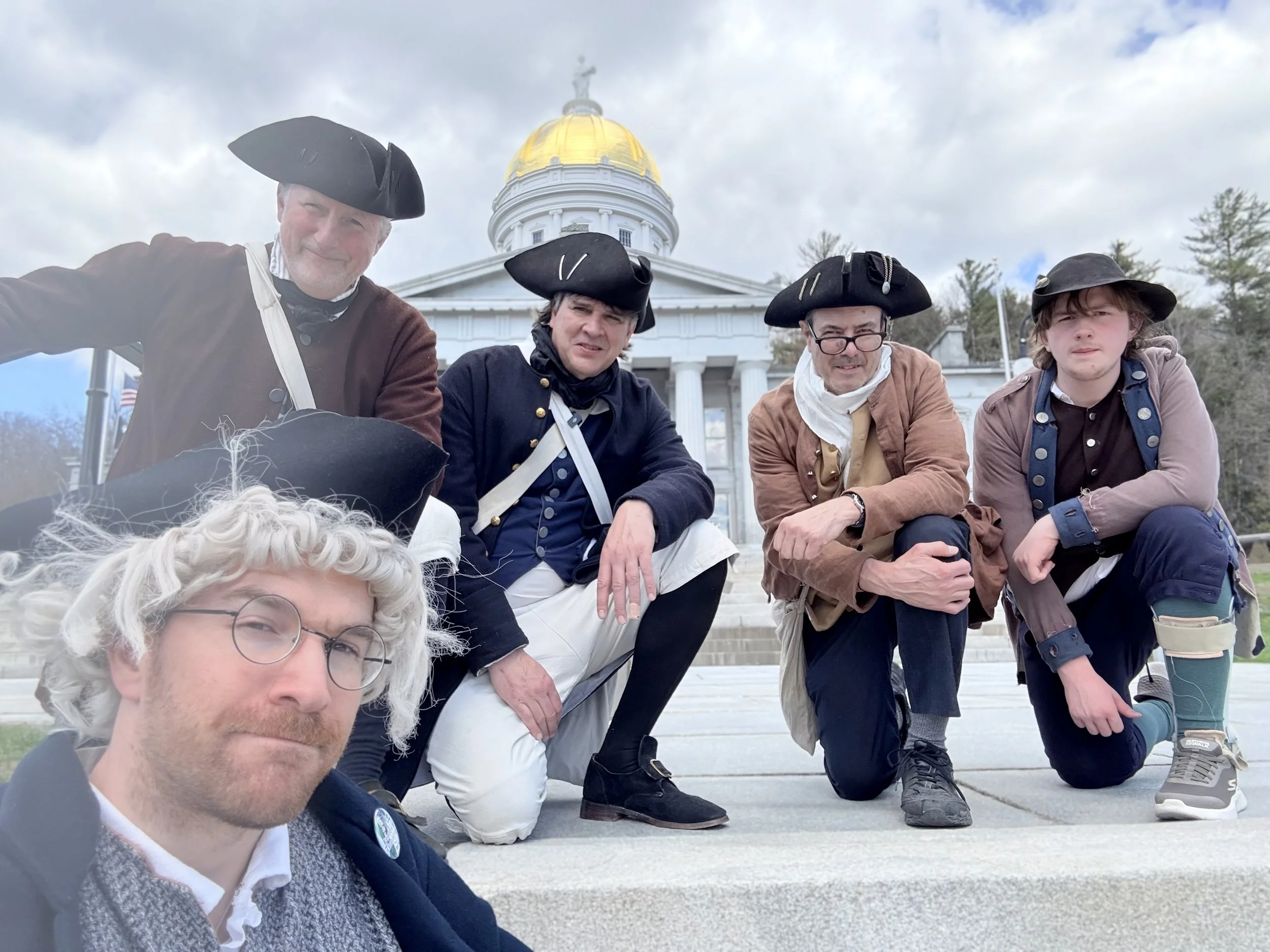 Four men dressed as Revolutionary War soldiers posing on steps in front of a historic white building with a gold dome, some smiling and wearing tricorn hats.