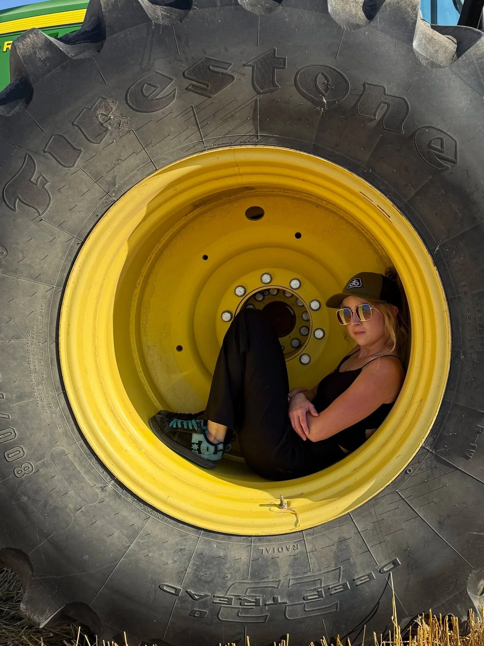 A person with blonde hair, wearing sunglasses, a baseball cap, and black clothing, sitting inside the hollow center of a gigantic tractor tire with yellow rim and black tread, outdoors with dry grass at the bottom.