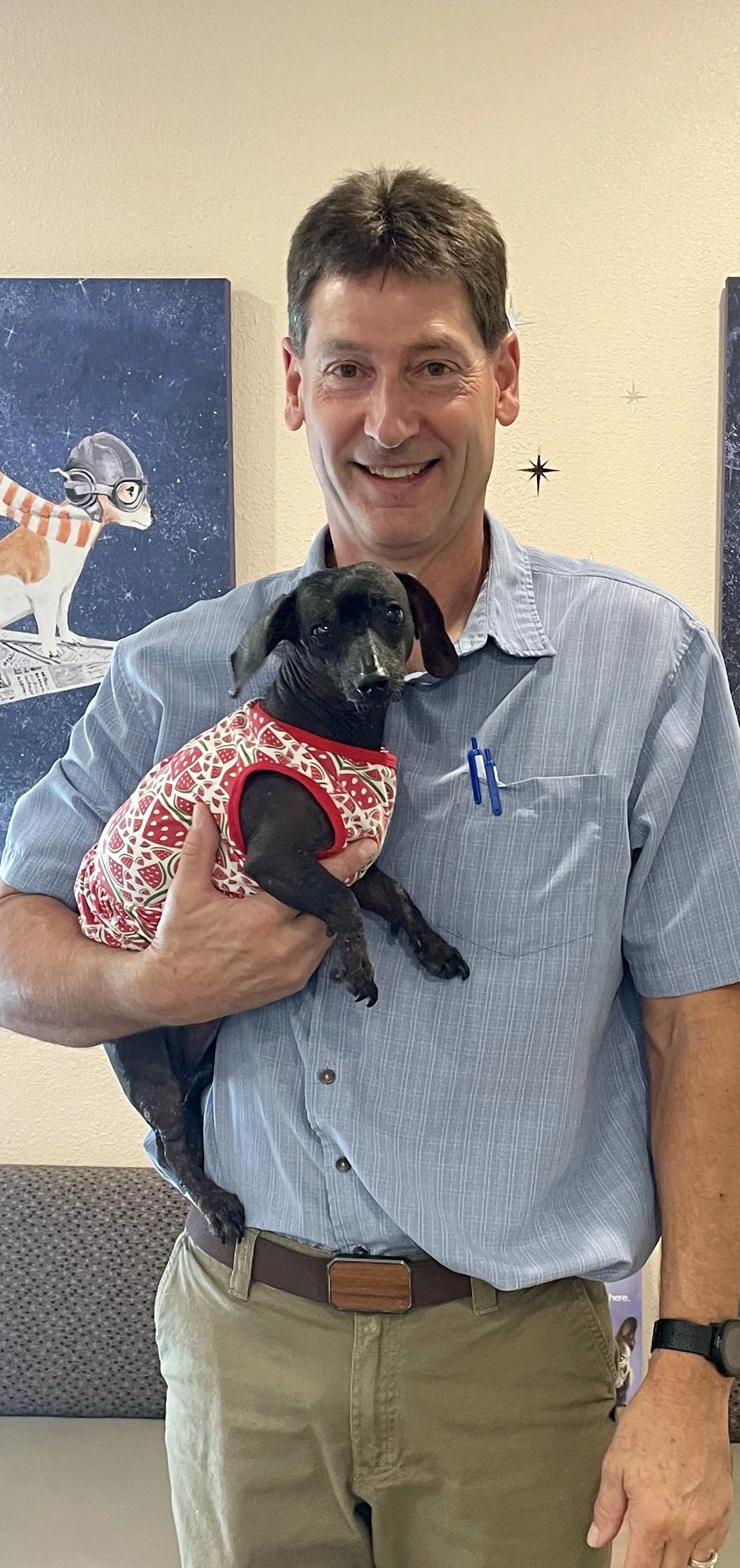 A man holding a small black dog wearing a red and white patterned shirt, standing inside a room with a yellow wall and space-themed artwork in the background.