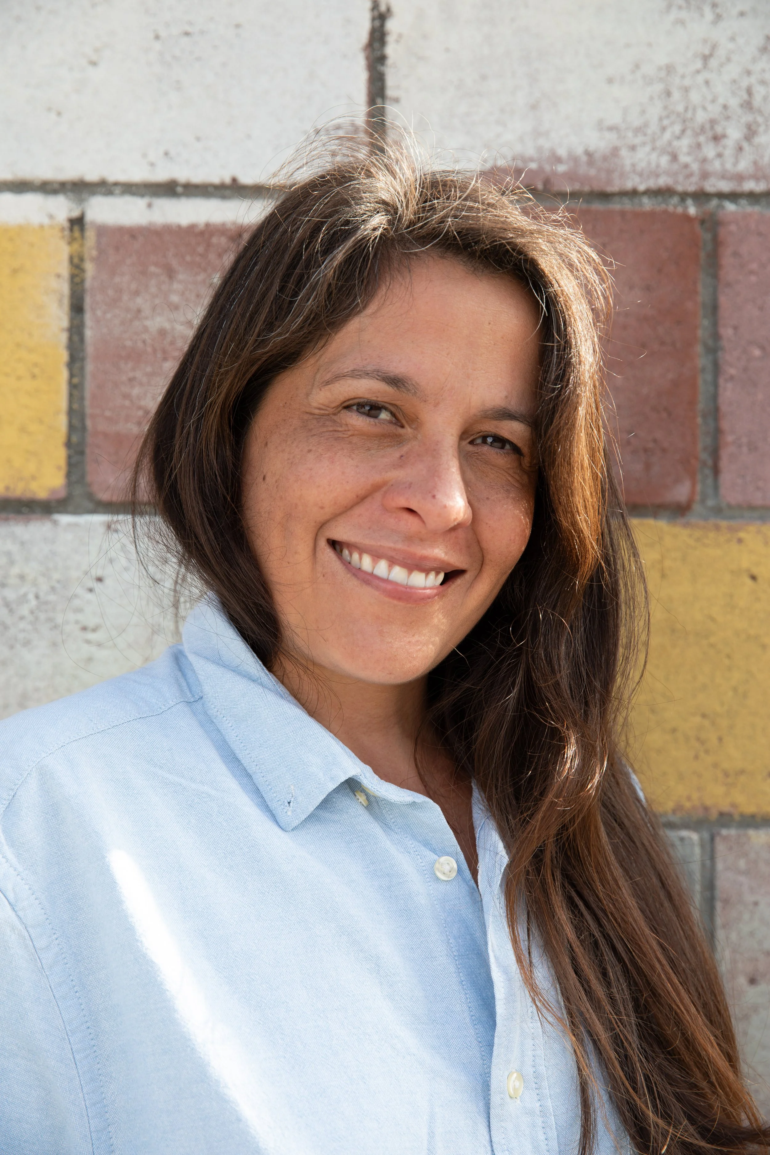Close-up of a smiling woman with long, wavy hair, wearing a light blue shirt, standing outdoors in front of a brick wall.