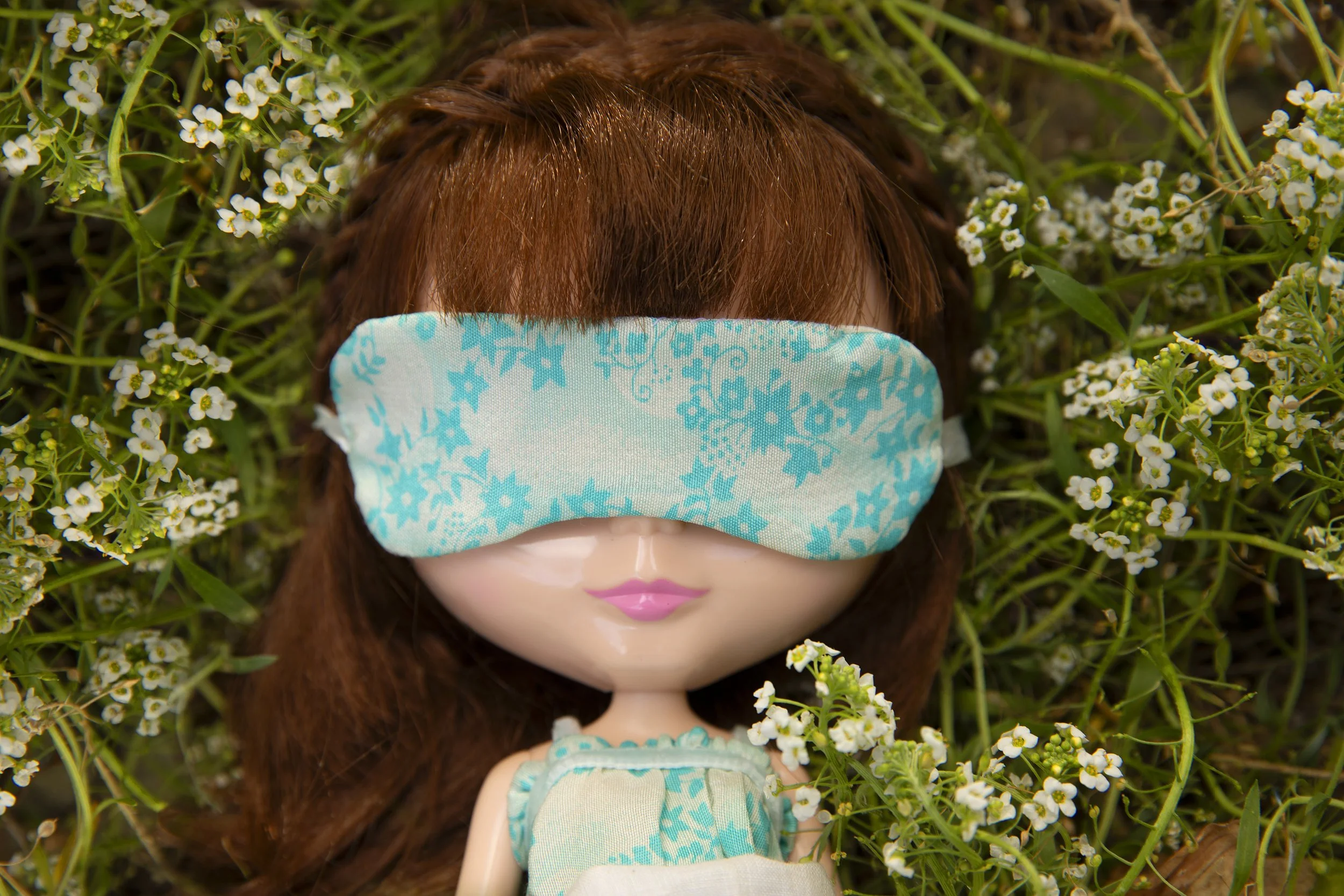 A doll with brown hair lying in a bed of small white flowers, wearing a blue and white dress and a matching eye mask.