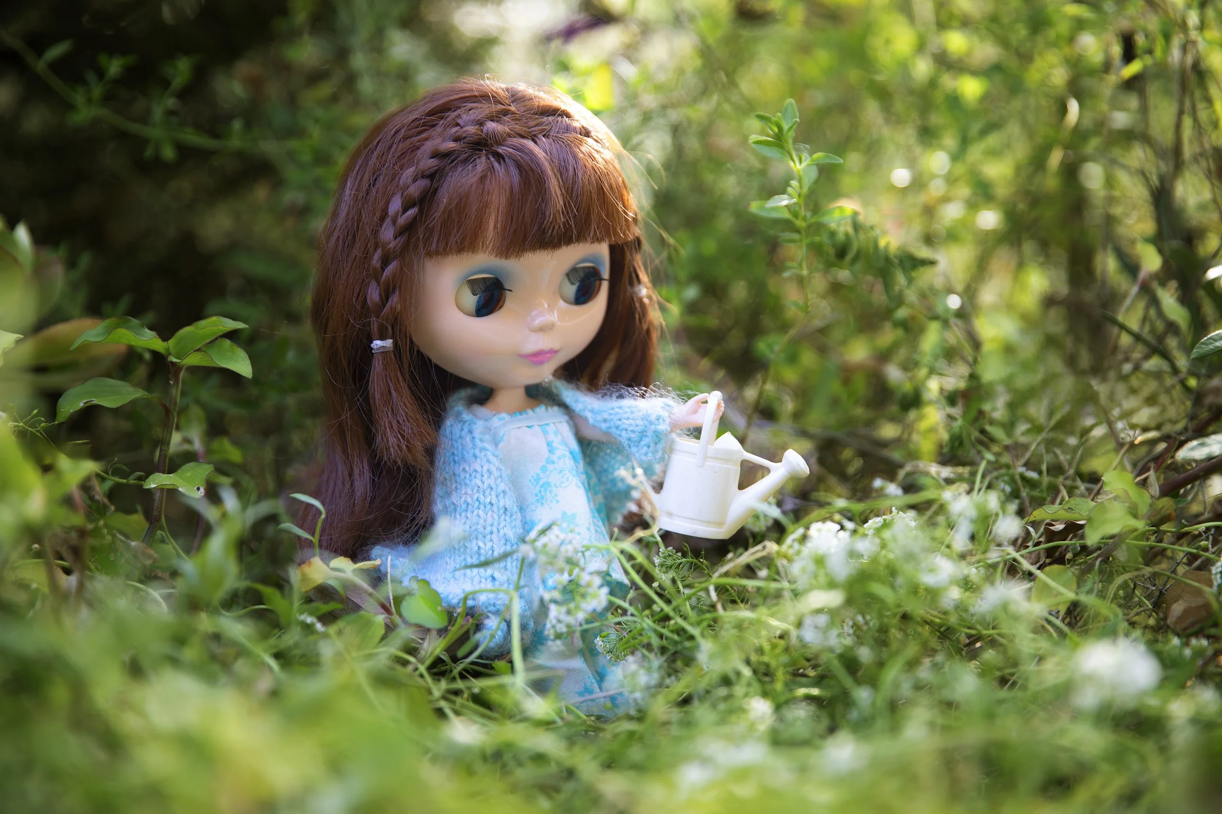 A doll with brown hair in braids and big eyes, dressed in a light blue outfit, sitting among green bushes and holding a white cup.