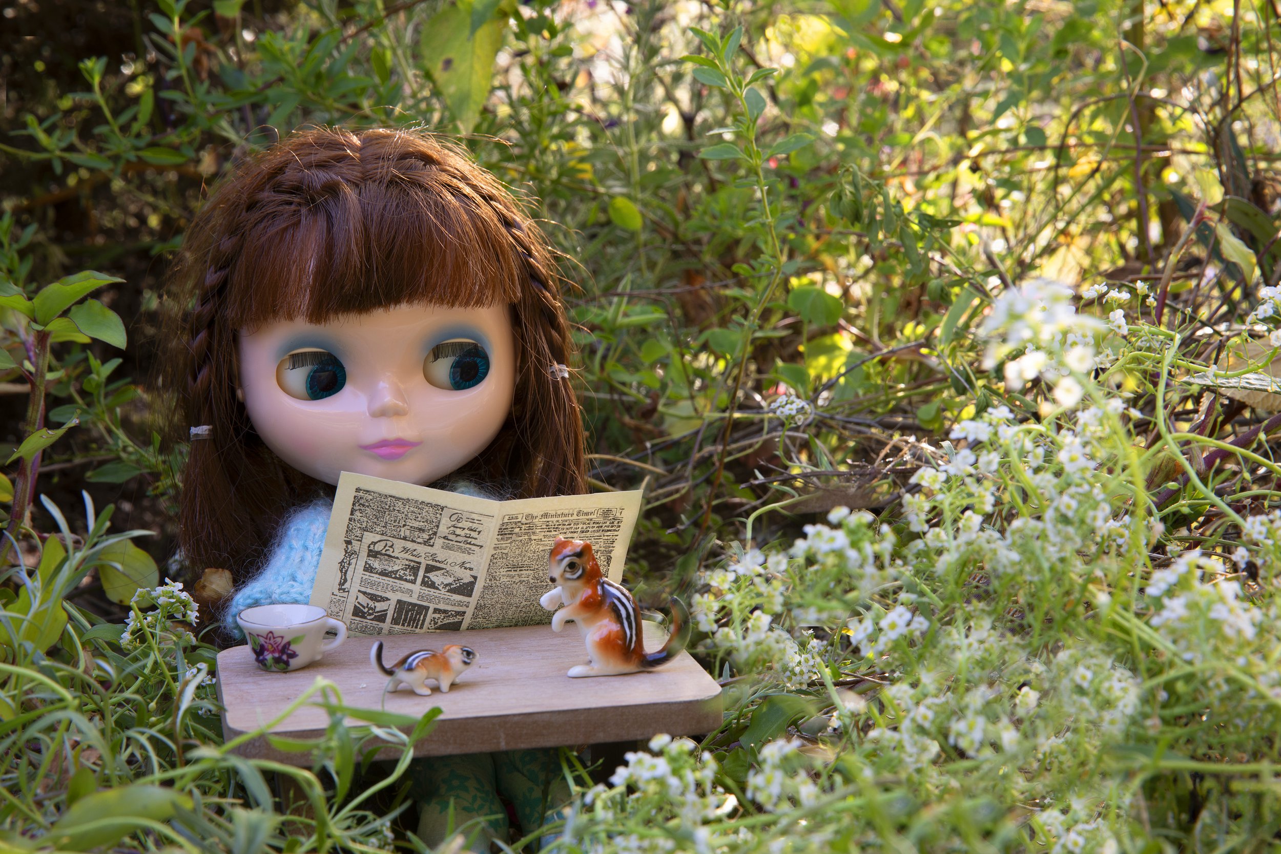 A doll with brown hair and large blue eyes sits among green plants and white flowers, reading a newspaper. There are small toy animals and tea cups on a wooden surface in front of her.