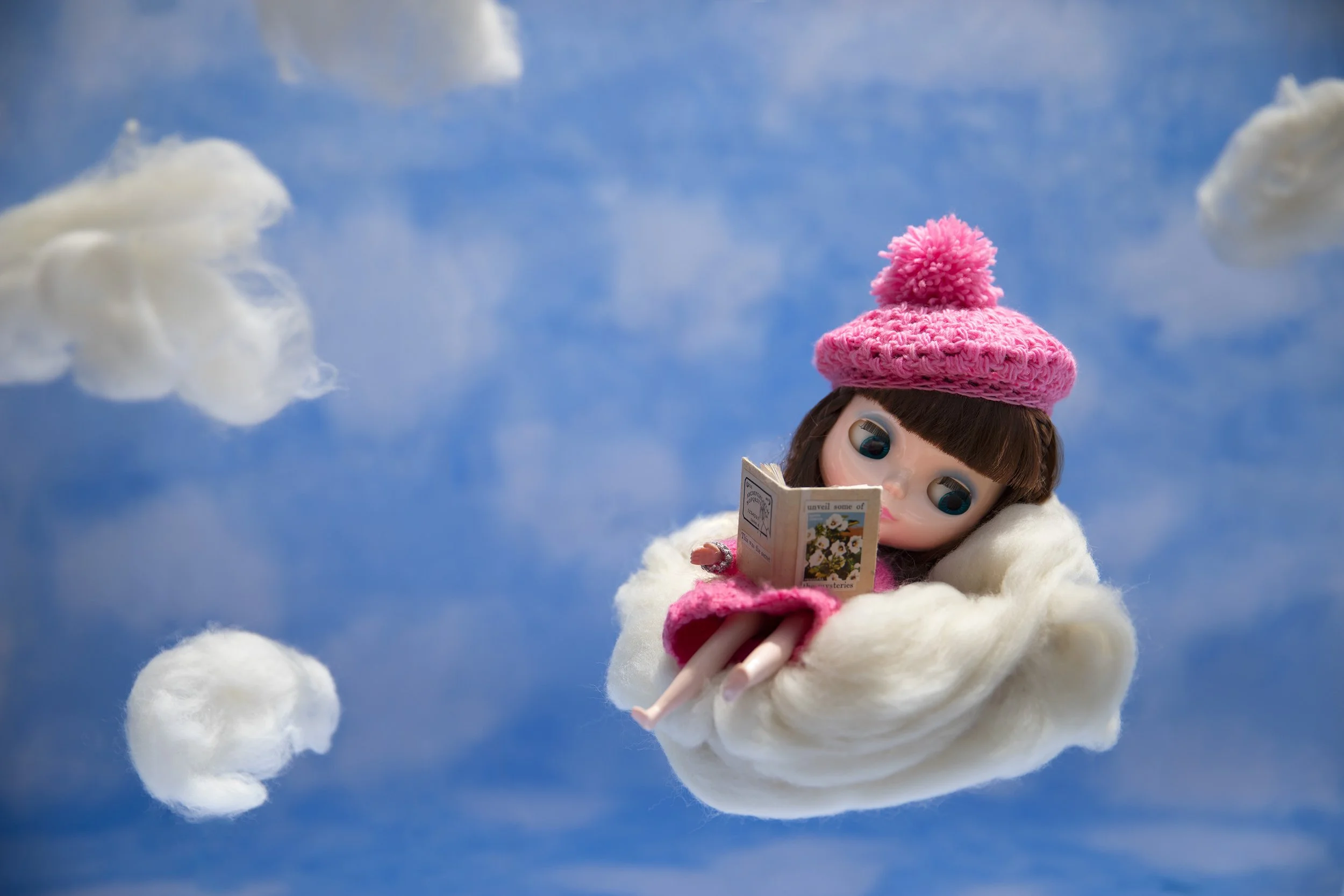 A doll with large eyes reading a book while sitting on a fluffy cloud in a sky with clouds.