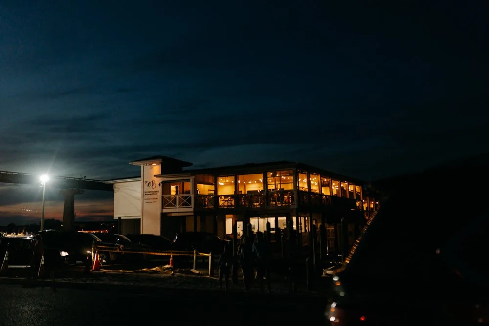 A two-story building with large windows illuminated from inside, set against a dark night sky. There are parked cars in front and a few people near the entrance.