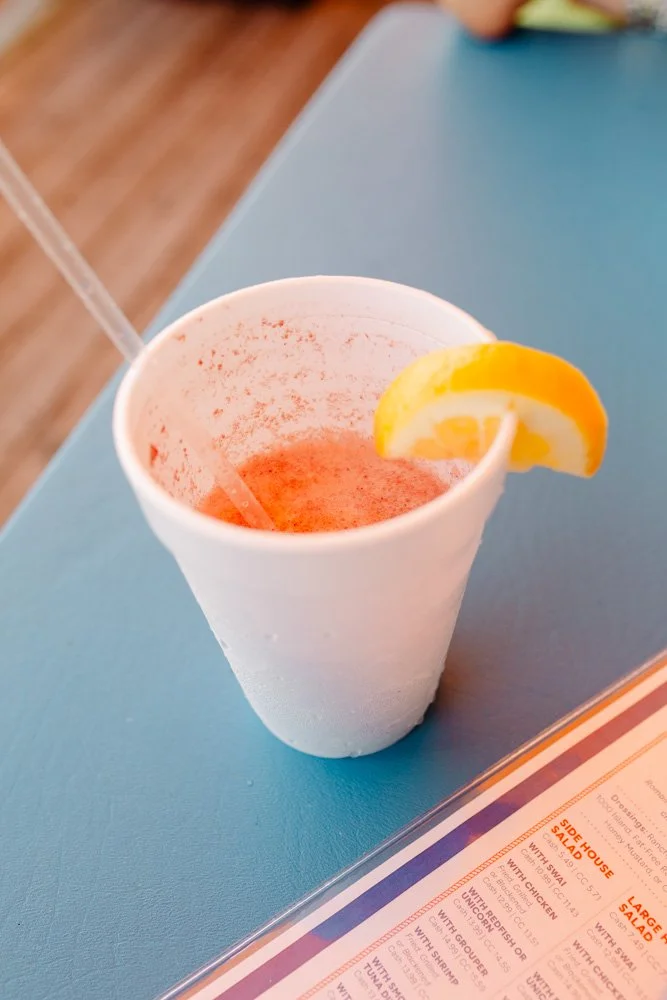 A white paper cup containing a pink cocktail with a lemon wedge garnish, placed on a blue table.