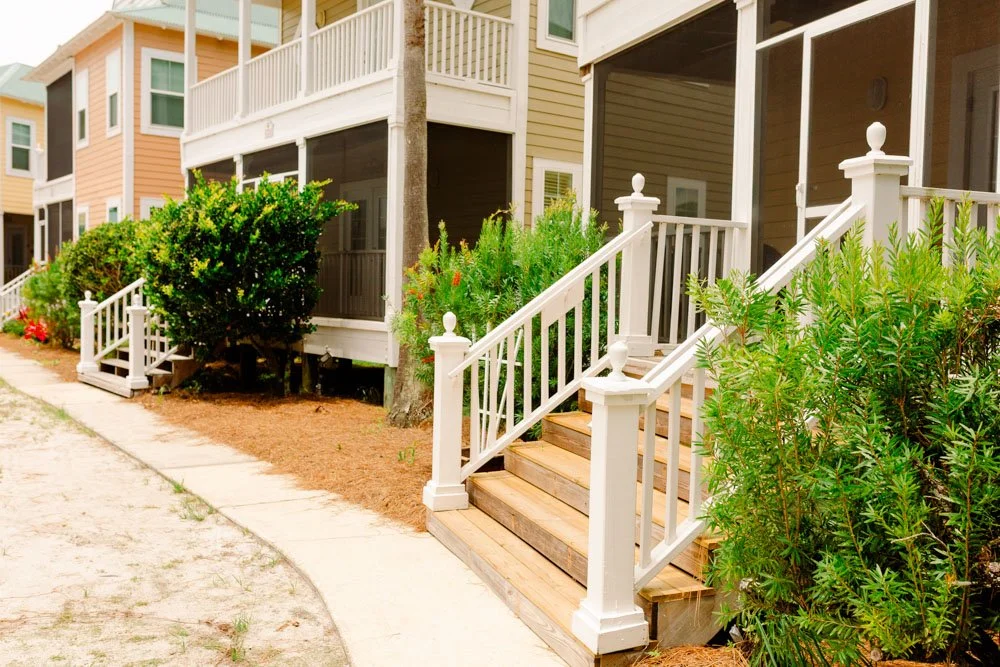 Front view of colorful suburban townhouses with white porches and stairs, surrounded by bushes and trees.