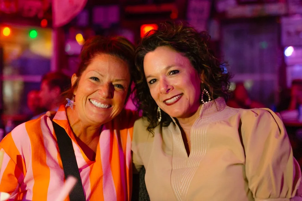 Two women smiling at a social event with colorful lighting and other people in the background.