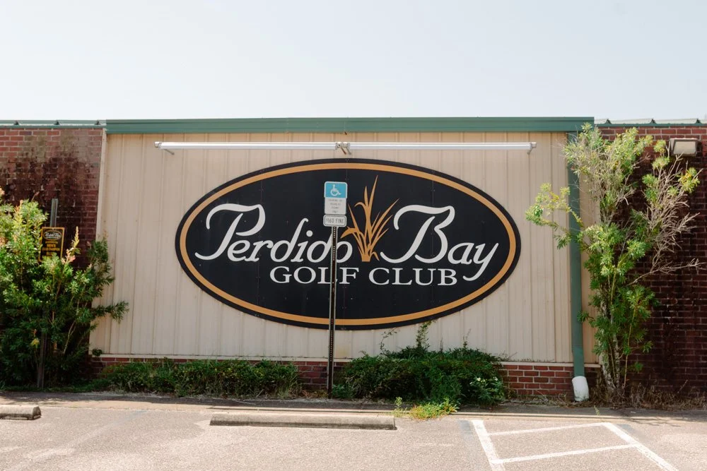 Sign for Perdido Bay Golf Club on a beige and brick building with some greenery and a handicapped parking sign in front.