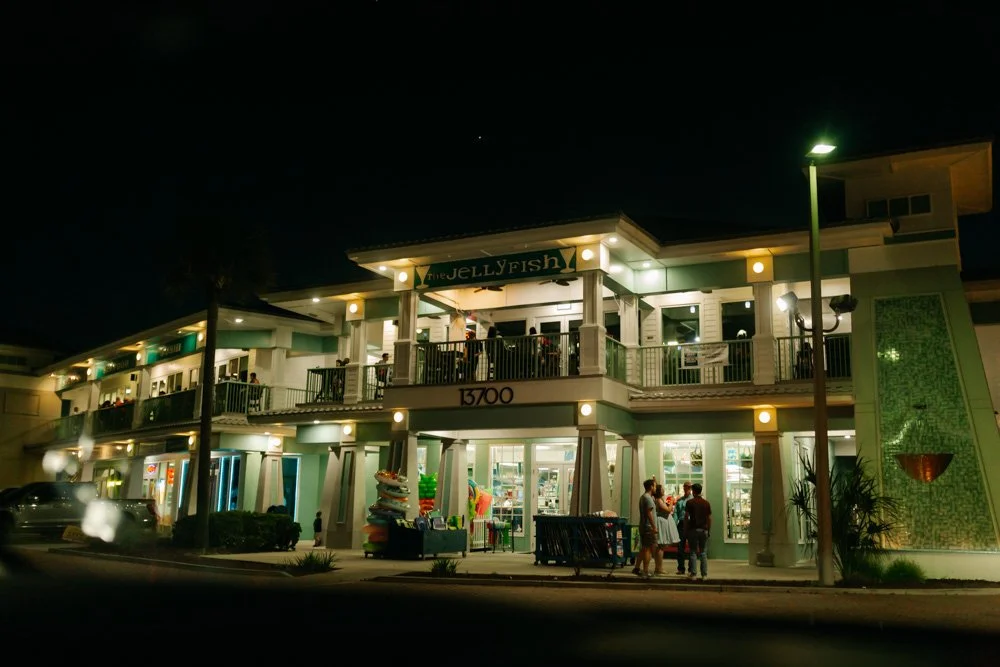 Night view of a two-story building with a sign that reads 'The Jellyfish', illuminated by lights. People are gathered outside, and the building has large windows showing inside stores. There are streetlights and a few parked cars nearby.