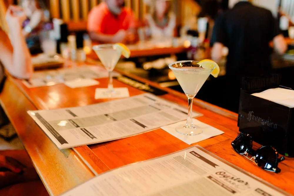 Two cocktails with lemon slices on a bar counter, with menus and sunglasses nearby, and people in the background.