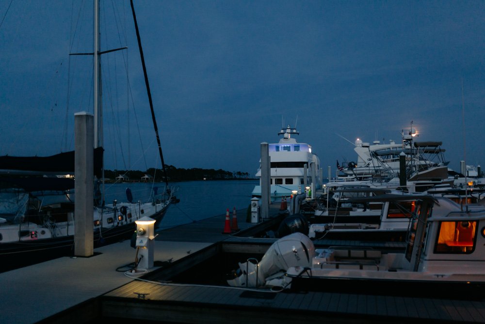 Boats docked at a marina during dusk, with some boat lights on and a calm water surface.