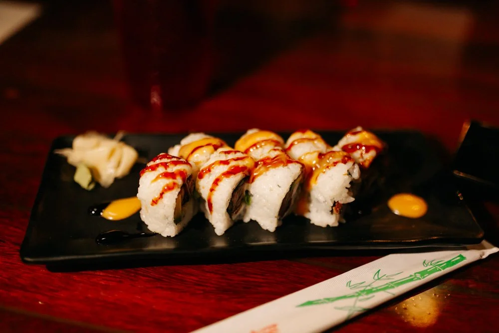 Sushi rolls topped with sauce on a black rectangular plate, accompanied by wasabi and pickled ginger, placed on a dark wooden table.