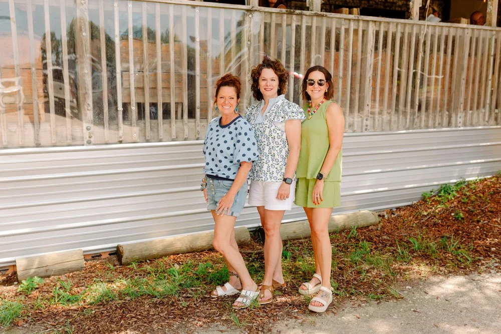 Three women standing outdoors in front of a metal and glass railing, smiling at the camera. They are dressed in casual summer clothing and are standing on a patch of dirt with some greenery.