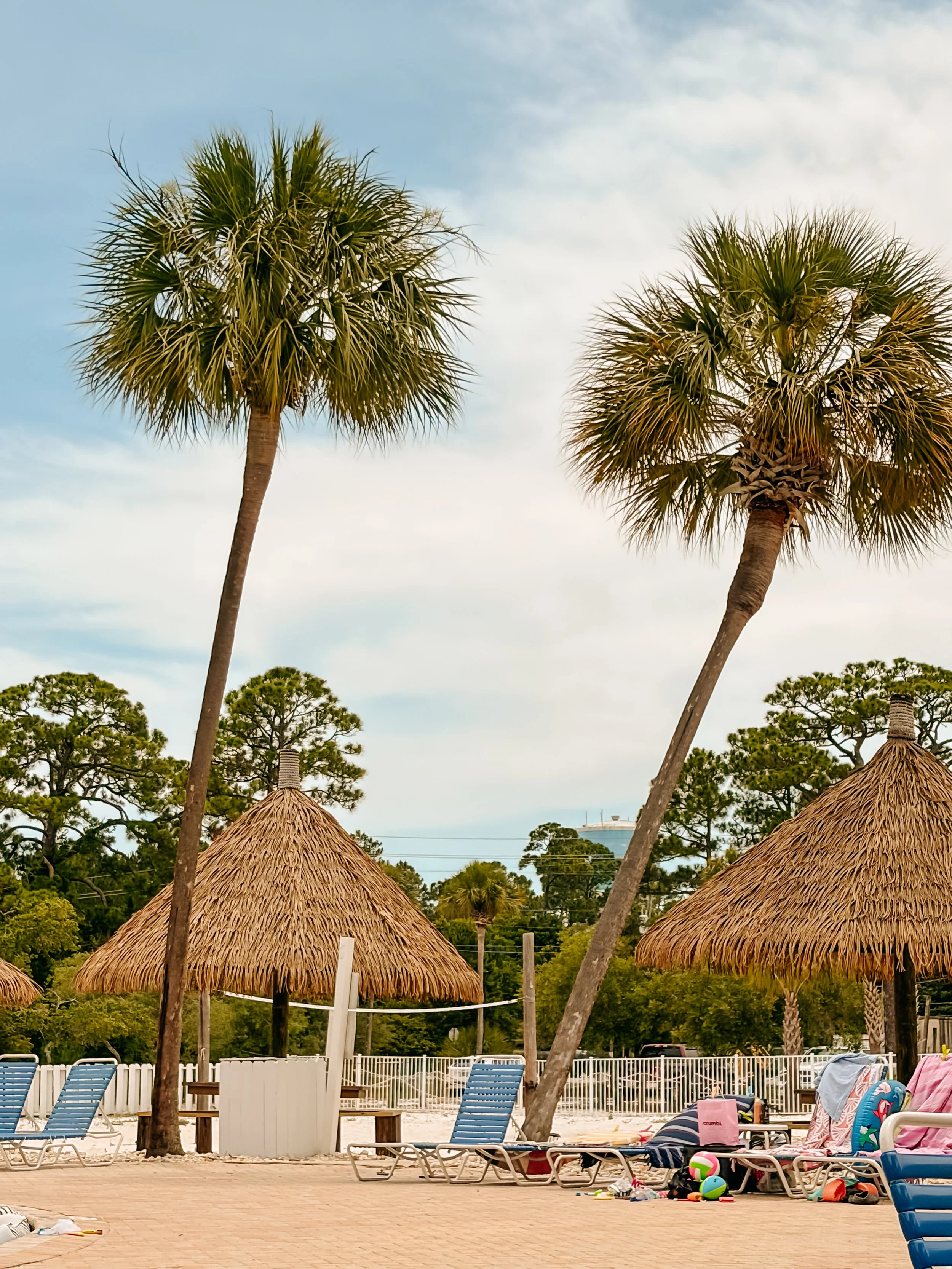 Poolside scene with lounge chairs, umbrellas, and toys, with trees and a cloudy sky in the background.