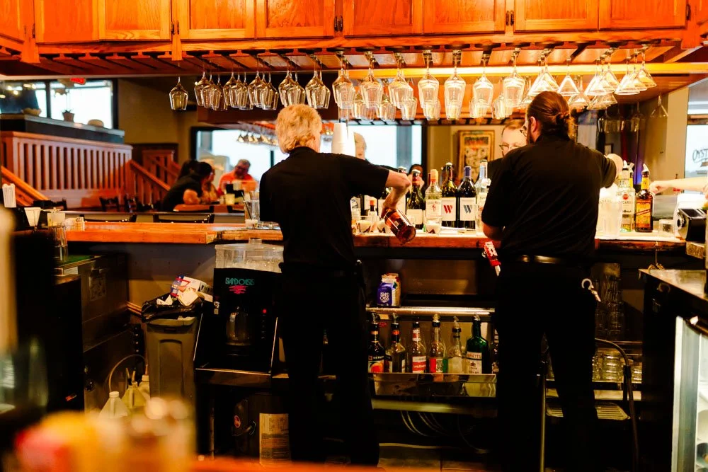 Bartenders preparing drinks at a bar counter in a restaurant with patrons in the background.