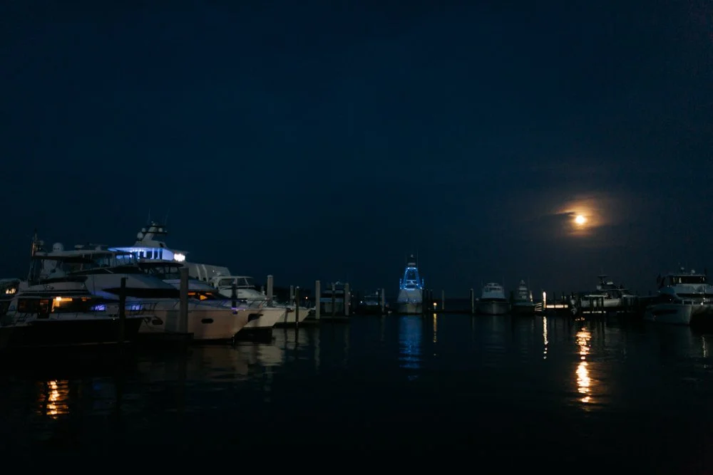 Nighttime scene of a marina with multiple boats docked, illuminated by underwater lights, under a dark sky with a bright moon.