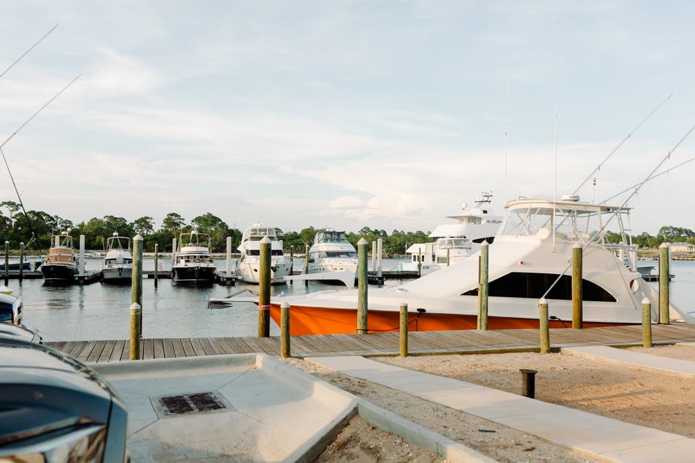 Marina with multiple docked boats, including a large white yacht on a wooden dock, under a partly cloudy sky.