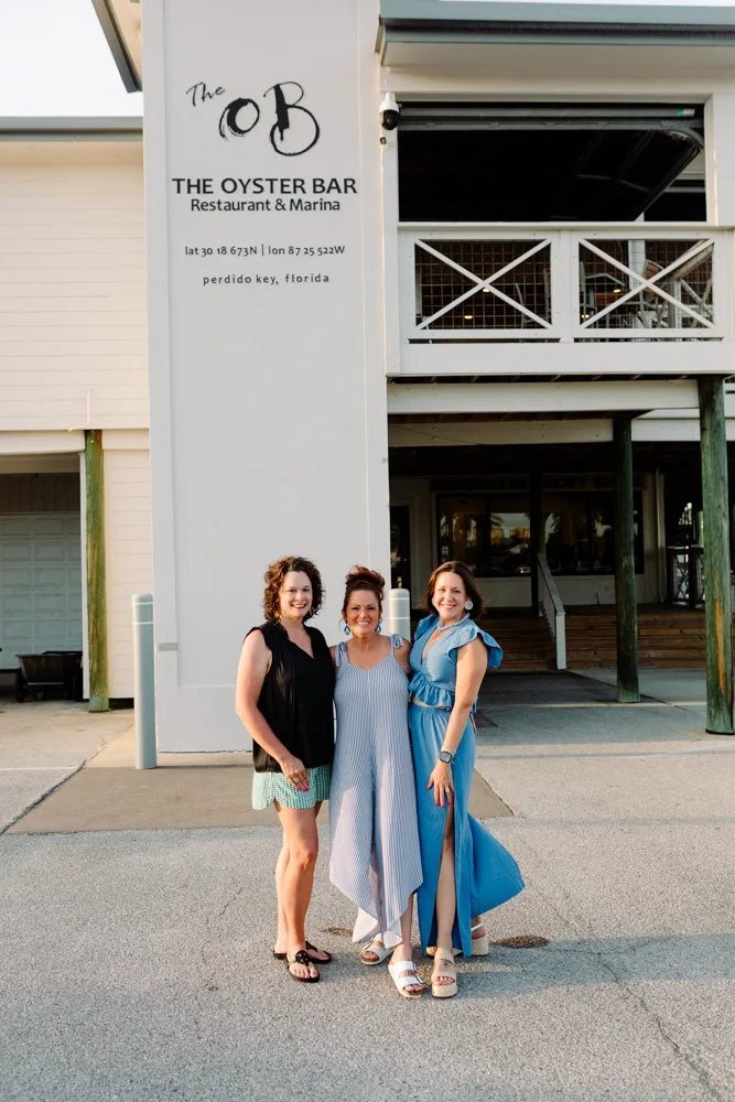 Three women standing outside the Oyster Bar restaurant and marina in Perdido Key, Florida, smiling at the camera during the daytime.