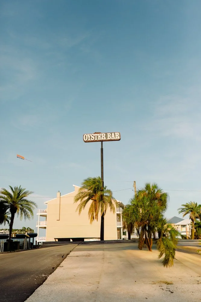 A building with a sign that reads 'Oyster Bar' and several palm trees in front, under a blue sky.