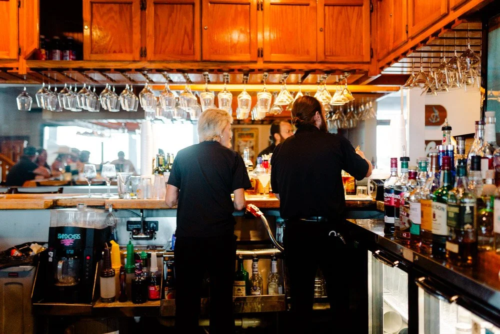 Two bartenders working behind a bar with liquor bottles and hanging glasses in a cozy restaurant or pub.