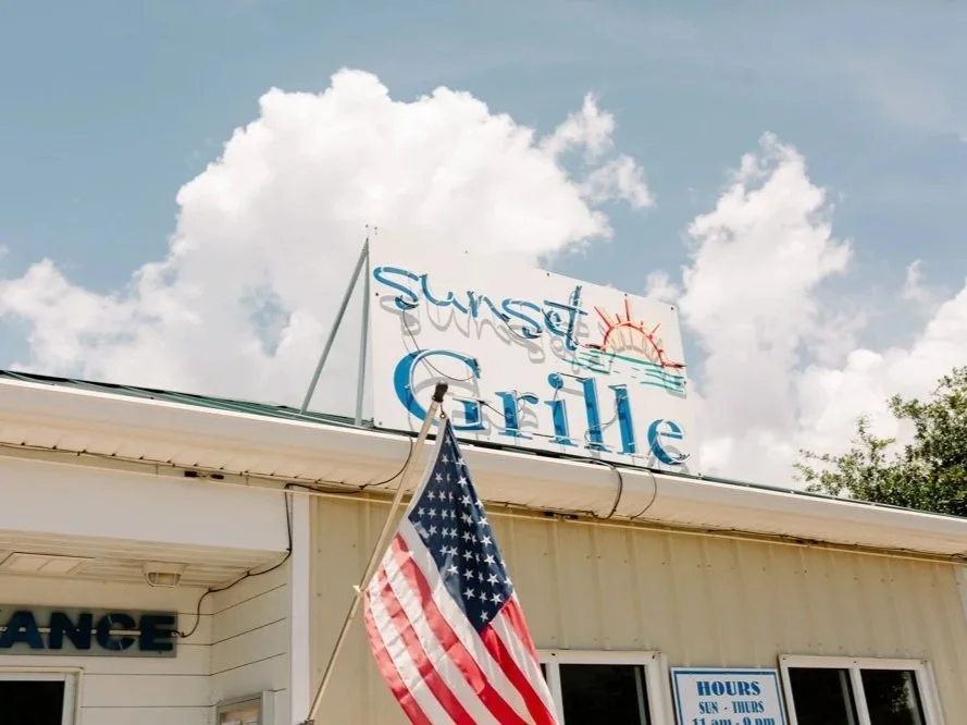 A building with a sign that reads "Sunset Grille" decorated with a drawing of a sunset, and an American flag in front of it.
