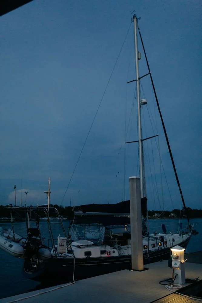 Sailboat docked at a marina during twilight, tied to a post with nearby electric outlet and water connection.