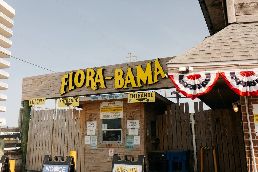 The entrance of Flora-Bama, a building with large yellow sign reading 'FLORA-BAMA', decorated with red, white, and blue patriotic bunting, featuring booth windows with signs for entrance and exit, and a wooden fence.