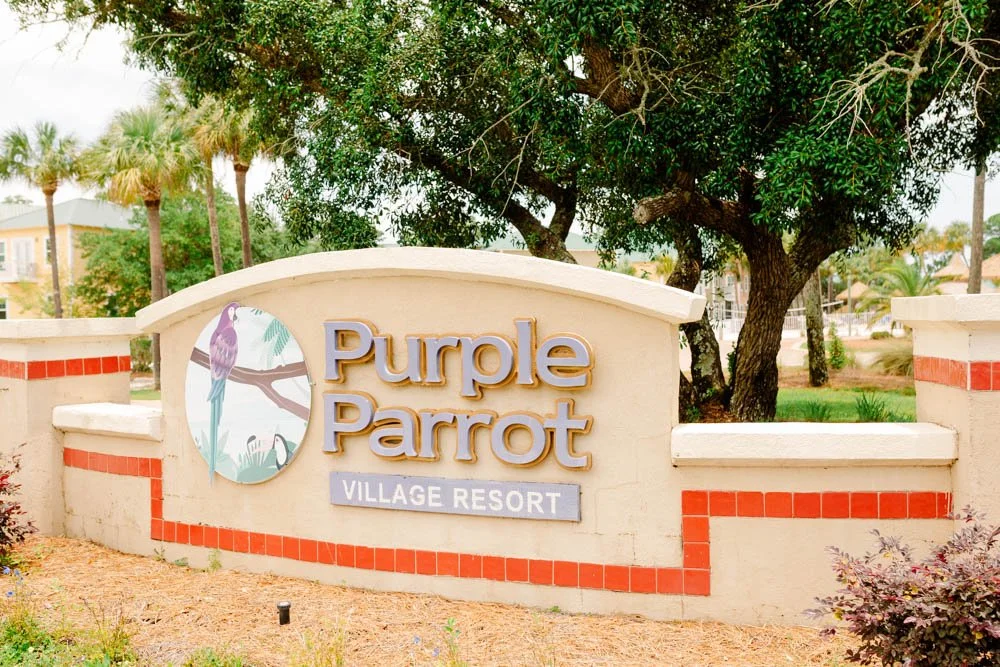 Sign for Purple Parrot Village Resort with a purple parrot illustration, surrounded by trees and plants.