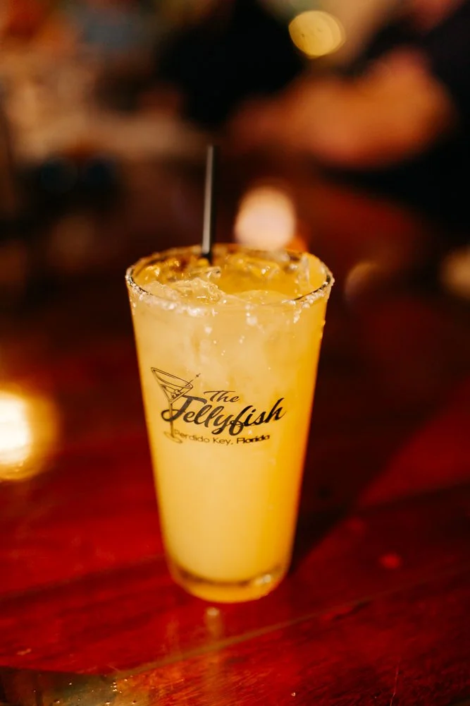 A yellow cocktail with ice in a tall glass. The glass has a logo that says 'The Jellyfish' and 'Orlando Key, Florida'. The rim is salted or sugared. A black straw is in the drink.