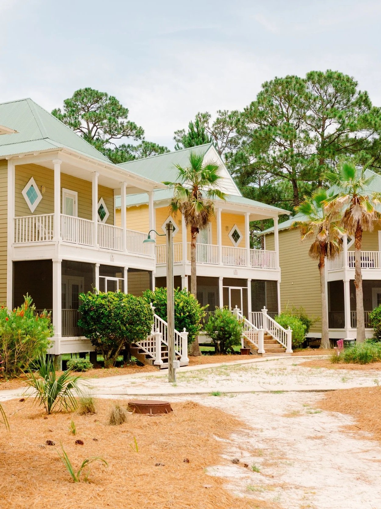 Colorful multi-story houses with white balconies and steps, surrounded by palm trees, bushes, and pine trees, under a cloudy sky.