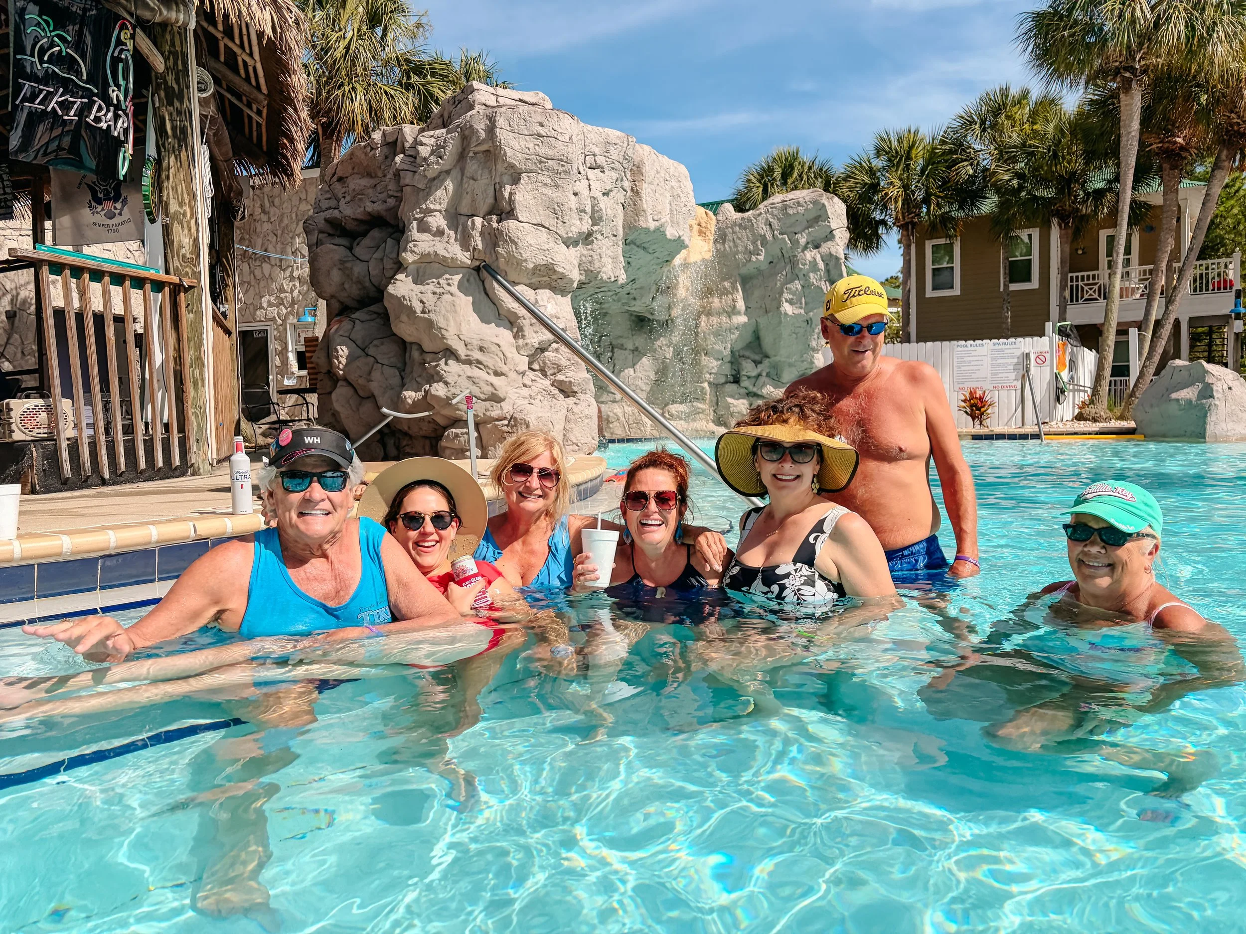 A group of smiling people enjoying time in a swimming pool with a water feature and rock formation in the background. Some wear sunglasses and hats, and a man is shirtless wearing a yellow cap. There are trees, residential buildings, and signs visibl