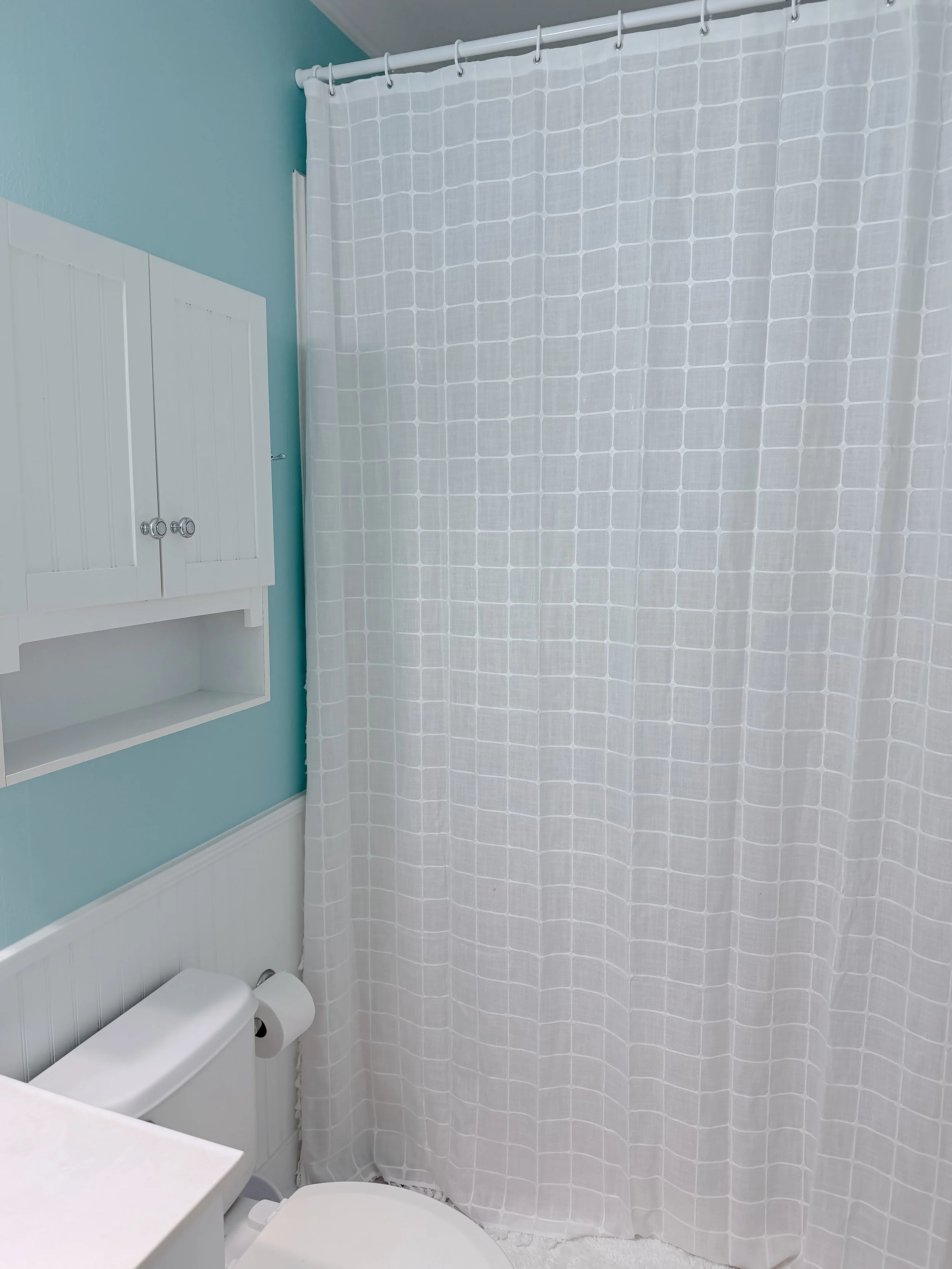 Bathroom with light blue walls, white bathroom cabinet, white toilet paper roll holder with toilet paper, white patterned shower curtain, and white beadboard wainscoting.