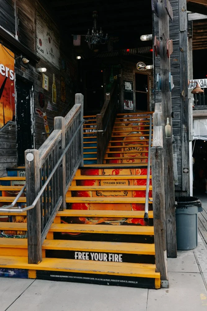 Wooden staircase with yellow steps and orange handrails, leading to an entrance in a rustic bar or restaurant, with various neon signs and posters on the dark wooden walls.