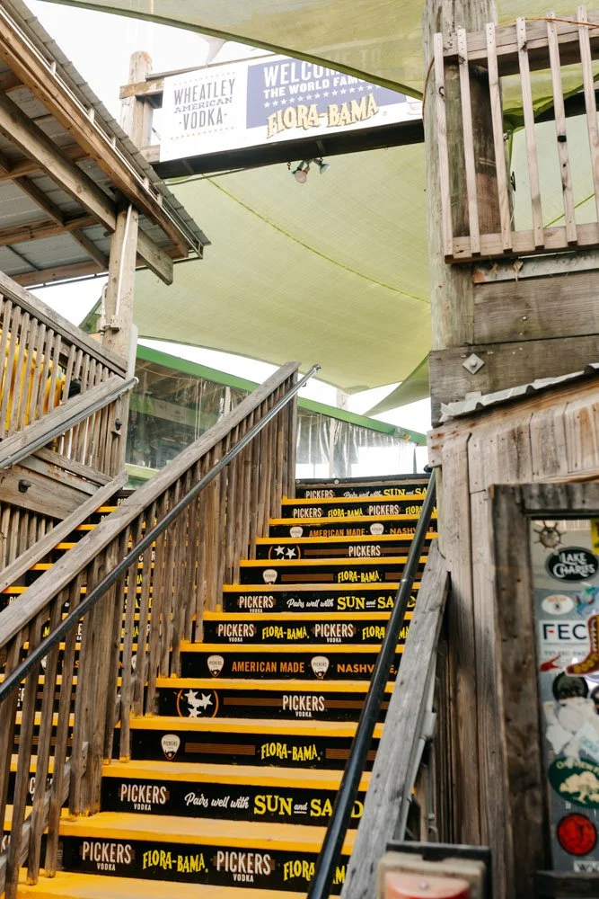 Outdoor wooden staircase with black and yellow steps advertising Pickers Vodka, Flora-Bama, and Sun, with signage and part of a building structure visible outside.