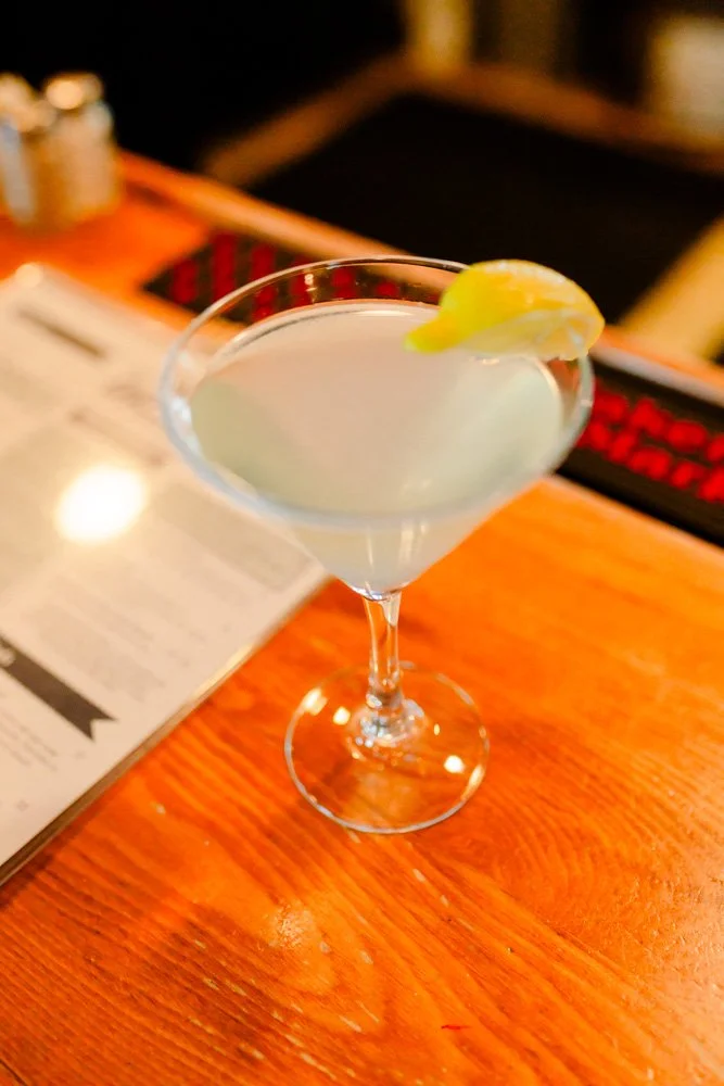 A cocktail glass with a light-colored drink garnished with a lemon wedge on the rim, placed on a wooden bar counter.