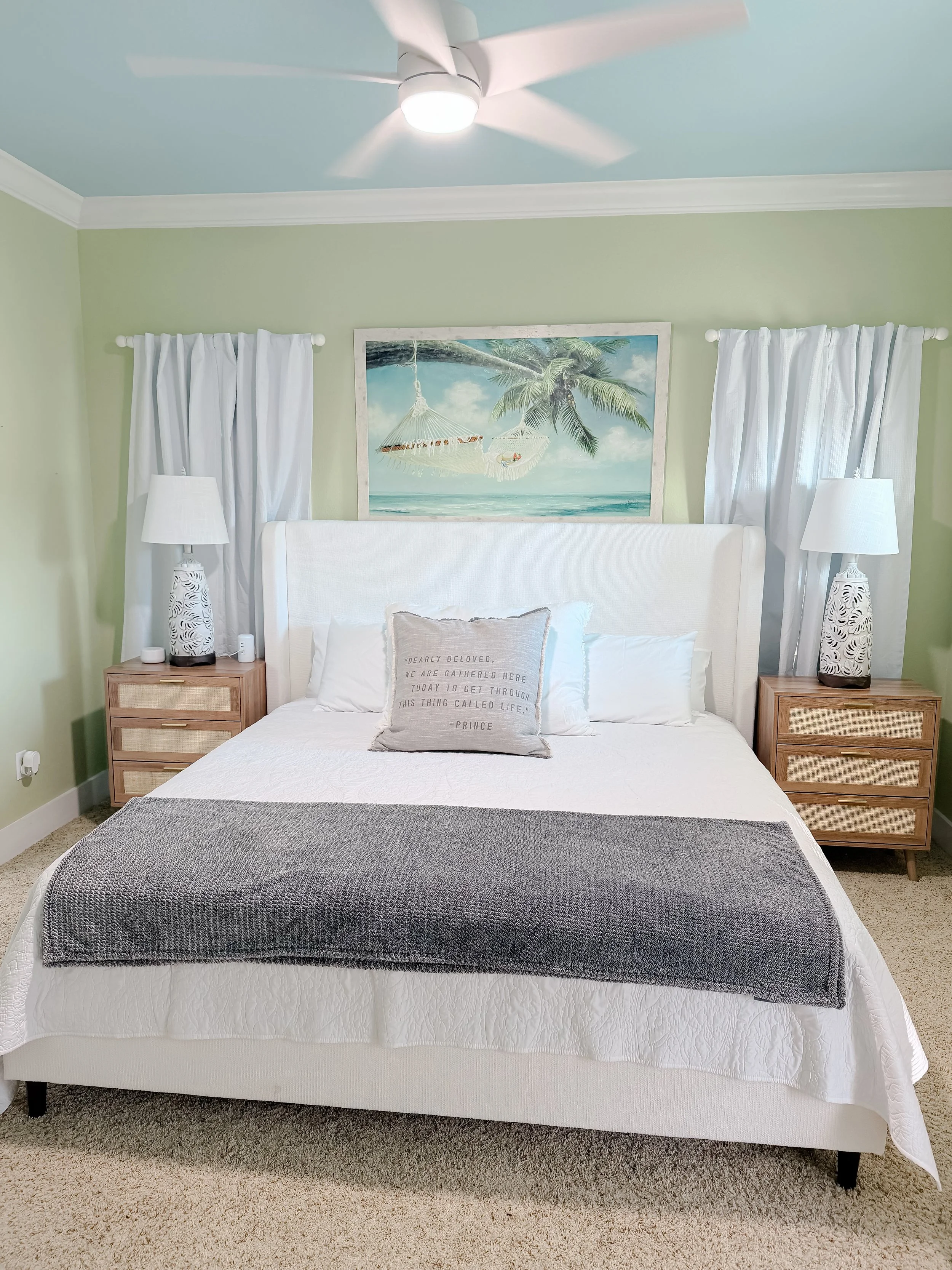 A bedroom with a white bed, gray blanket, and decorative pillow with quote, flanked by two wooden nightstands with white lamps, wall art of a beach scene, green walls, and white curtains.