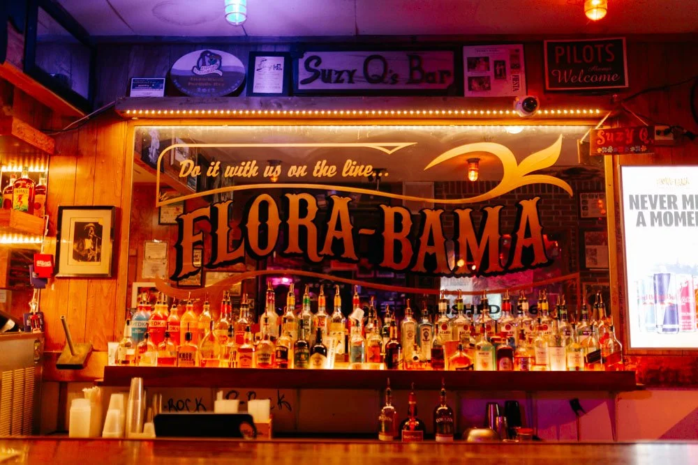 Bar with illuminated shelves filled with bottles, a large sign reading 'Flora-Bama', and various signs and decorations on wood-paneled walls.